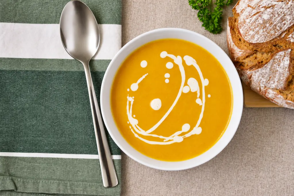 Overhead view of creamy pumpkin soup with a spoon, bread and green cloth